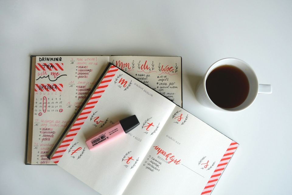 An open planner with pink hand lettering, a pink highlighter and a cup of tea on a white desk