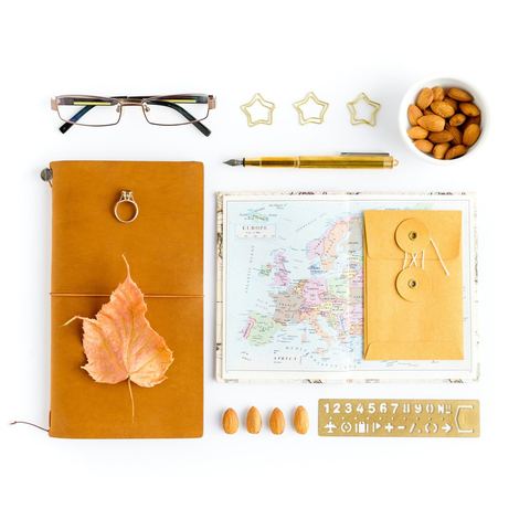 A leather travel journal with a pressed leaf, a map of Europe, glasses and a pen laid out on white