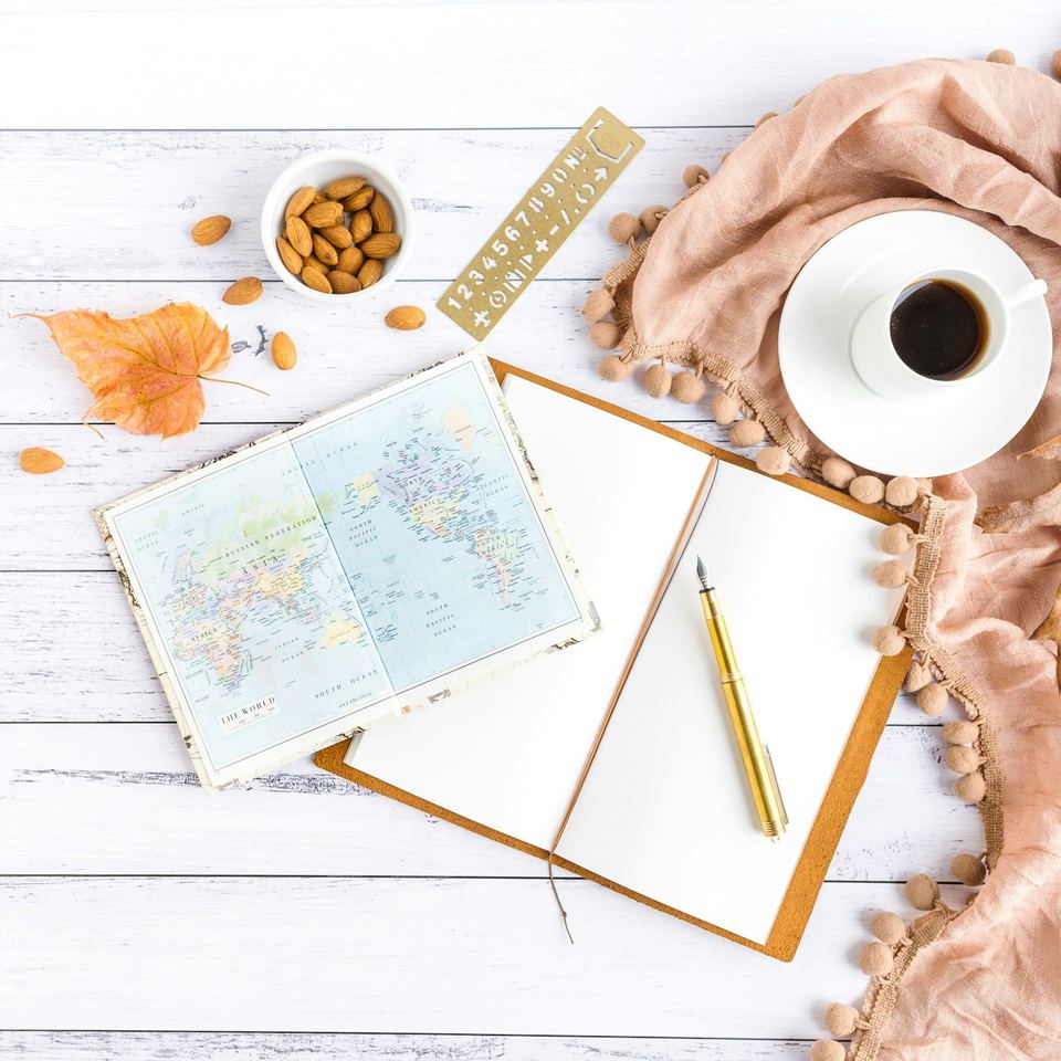 A folded map, an open notebook and a cup of coffee on a white table beside a blush-pink throw